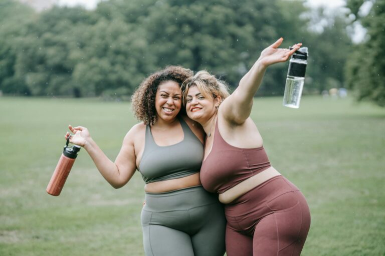 Two women in sportswear smiling and hugging outdoors, holding water bottles.